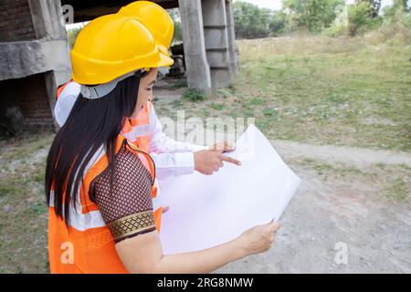 Two Indian male and female civil engineers or architect wearing helmet and vest holding paperwork blueprint at construction site discussing real estat Stock Photo