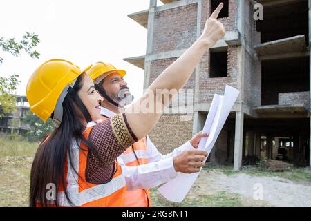 Two Indian male and female civil engineers or architect wearing helmet and vest holding paperwork blueprint at construction site discussing real estat Stock Photo
