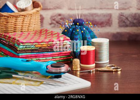 Stack of colorful fabrics on table surrounded by sewing accessories on brick wall background Stock Photo