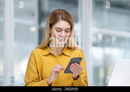 Mature experienced business woman inside office at workplace, employee sitting at desk smiling holding phone in hand, boss using application on smartphone, browsing internet pages. Stock Photo