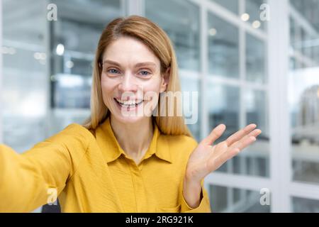 Online video call, businesswoman inside office at workplace looking at smartphone camera using video call app talking with friends and smiling joyfully. Stock Photo