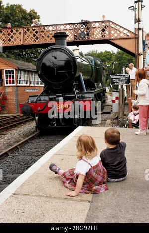 Two children sit on the platform at Buckfastleigh as the ex-GWR steam locomotive 3803 makes its way into the station.  The line is run by the South Devon Railway. Stock Photo