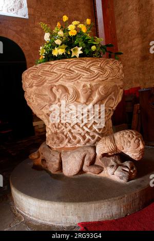 Norman font in St Michael and All Angels Church, Castle Frome, Herefordshire.  It is in the form of a large tub standing on three crouching men with beards (only one of which survives complete). Between guilloche borders can be seen the angel, eagle, lion and cow symbols of the four apostles; the central scene is a portrayal of Christ's baptism by John the Baptist. Stock Photo