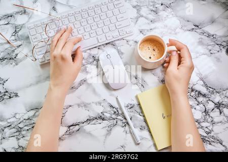 Flat lay, office desk with Girl holds coffee. Keyboard, supplies, cup of coffee on marble background. Stock Photo