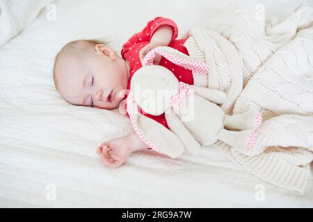 Baby girl sleeping under knitted blanket with her favorite toy. Little child in pink clothes having a day nap. Infant kid in nursery Stock Photo