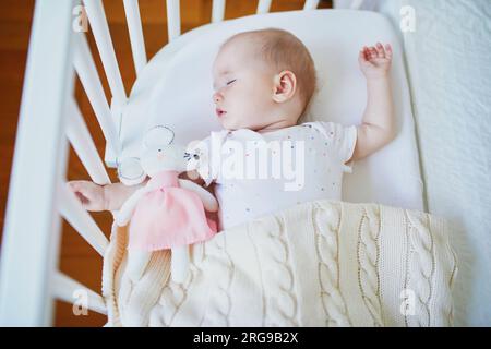 Adorable baby girl sleeping in co-sleeper crib attached to parents' bed with stuffed toy. Little child having a day nap in cot. Sleep training concept Stock Photo