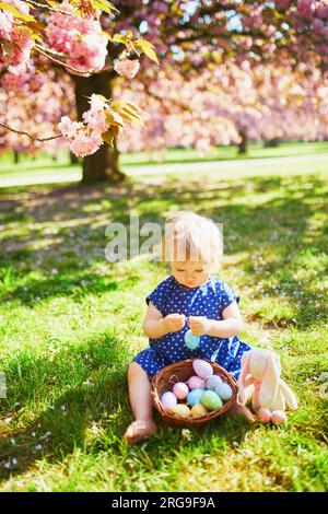 Cute little one year old girl playing egg hunt on Easter. Toddler sitting on the grass under cherry blossom tree in full bloom. Little kid celebrating Stock Photo
