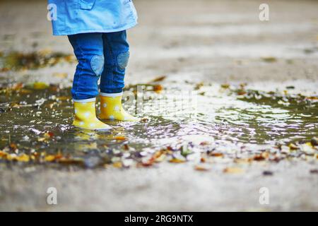 Child wearing yellow rain boots and jumping in puddle on a fall day. Toddler girl having fun with water and mud in park on a rainy day. Outdoor autumn Stock Photo