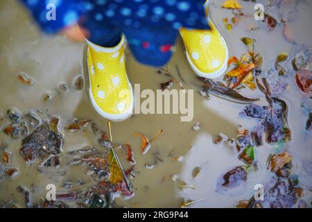 Child wearing yellow rain boots and jumping in dirty puddle on a fall day. Toddler girl having fun with water and mud in park on a rainy day. Outdoor Stock Photo