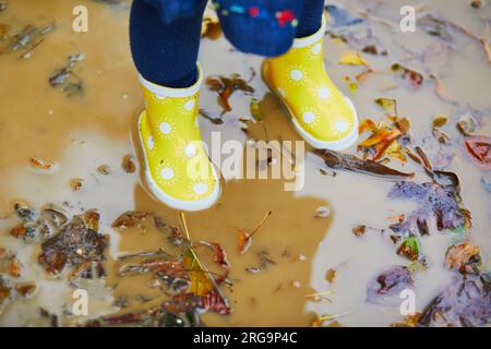 Child wearing yellow rain boots and jumping in dirty puddle on a fall day. Toddler girl having fun with water and mud in park on a rainy day. Outdoor Stock Photo