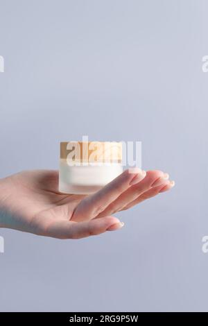 Hand holding blank white matte glass cream jar with bamboo lid on gray background. Cosmetic beauty product branding mockup. Skincare Stock Photo