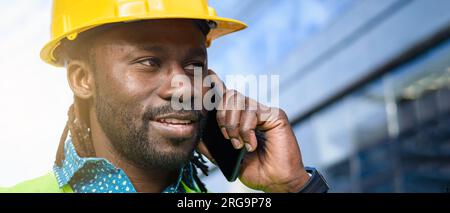 closeup banner of black engineer man with beard, dreadlocks and is wearing a yellow safety helmet, he is talking on the phone smiling happy hearing go Stock Photo