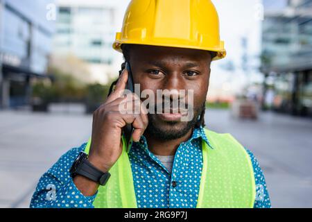 portrait of black engineer man using phone outdoors, on phone call, standing happy looking at camera, people concept. Stock Photo