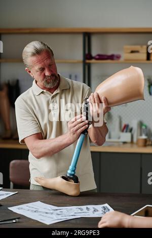 Vertical portrait of long haired senior craftsman building leg prosthetics in workshop and inspecting joints Stock Photo