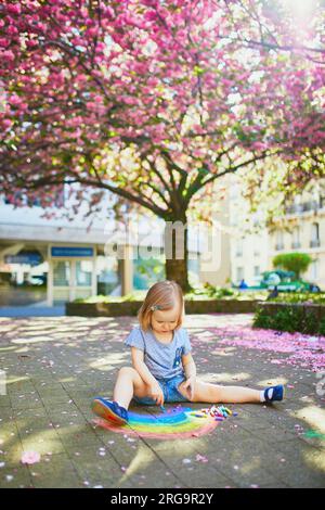Adorable toddler girl drawing rainbow with colorful chalks on asphalt in park with blooming cherry blossom trees. Outdoor activity and creative games Stock Photo
