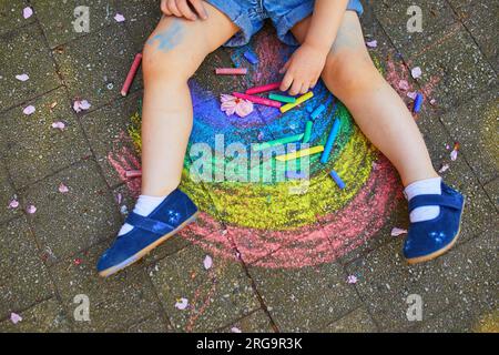 Toddler girl drawing rainbow with colorful chalks on asphalt. Outdoor activity and creative games for small kids Stock Photo