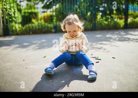 Adorable toddler girl at her dirty hands after drawing with colorful chalks on asphalt. Outdoor activity and creative games for small kids Stock Photo