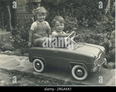 A twin brother and sister, wearing matching dungarees, play together in a splendid toy pedal car. Stock Photo