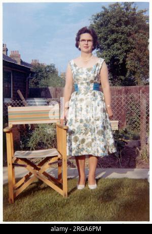 A middle aged woman in a floral summer dress standing somewhat stifly in a neat suburban back garden, one hand on a wood and canvas garden chair. Stock Photo