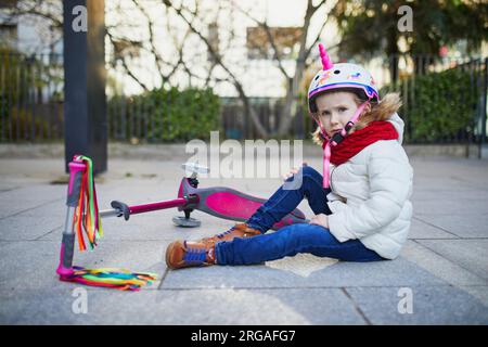 Preschooler girl in unicorn helmet sitting on the ground after she fell while riding her scooter in park on a spring day. Outdoor sport activities and Stock Photo