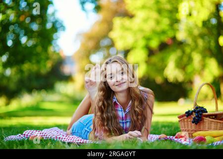 Girl having a picnic in park, lying on the grass, picnic basket with fruits near her Stock Photo