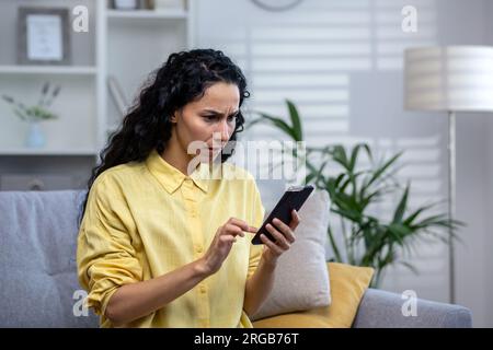 Upset and disappointed serious thinking woman alone at home using app on phone close up, hispanic sitting on sofa in living room at home. Stock Photo