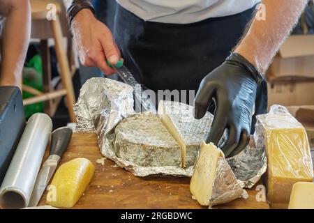 Seller's hands cut off a piece of cheese. Russian farmer's cheeses from the manufacturer. Assortment of tasty cheese on the counter. Stock Photo