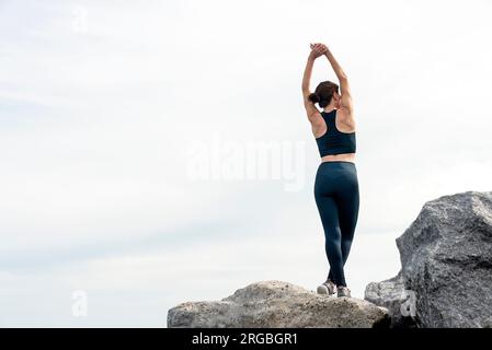 fit sporty woman doing a stretching exercise outside standing on rocks and boulders, rear view. Stock Photo