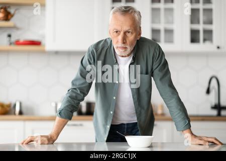 Portrait Of Pensive Senior Man Standing At Table In Kitchen Interior Stock Photo