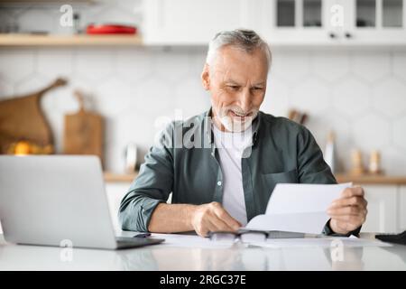 Happy Senior Man Reading Letter While Sitting At Desk In Kitchen Stock Photo