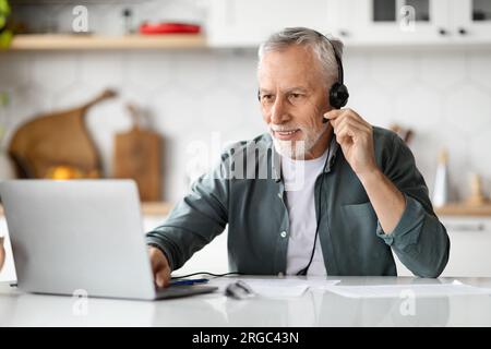Smiling senior man sitting in kitchen, using modern laptop and headset Stock Photo