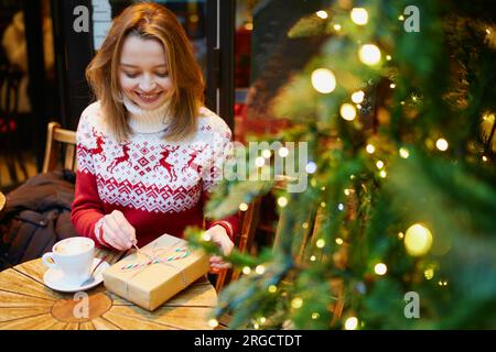 Cheerful young girl in warm red knitted holiday sweater drinking coffee or hot chocolate in cafe decorated for Christmas and unwrapping Christmas pres Stock Photo