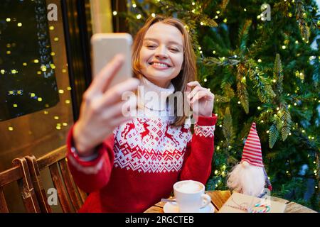 Cheerful young girl in holiday sweater in cafe decorated for Christmas, making selfie with her coffee cup and Christmas present Stock Photo