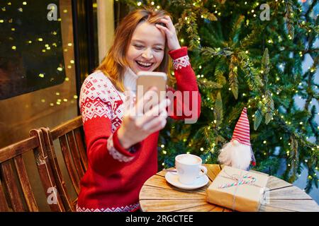 Cheerful young girl in warm red knitted holiday sweater in cafe decorated for Christmas, making selfie with her coffee cup and Christmas present Stock Photo