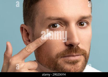 Face of serious bearded middle aged man applying face cream on cheek under eye looking at camera. Stock Photo