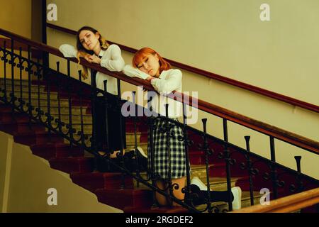 Two female students in uniforms are together on a university staircase, both appearing sad. One student is blonde, while the other is a ginger Asian. Stock Photo