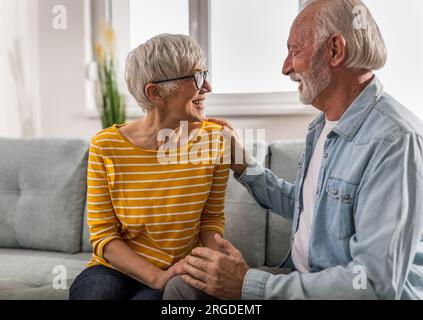 Senior couple sitting on sofa in living room talking and man holding his hand on woman's shoulder as support sign Stock Photo