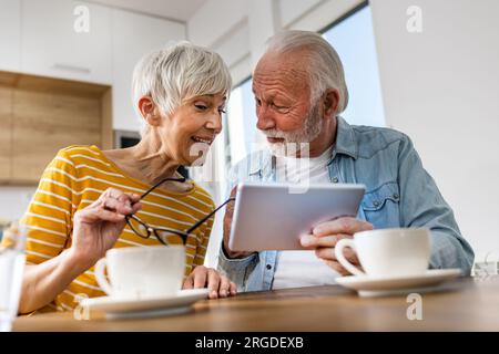 Senior couple sitting at dining table and looking at tablet, surfing internet Stock Photo