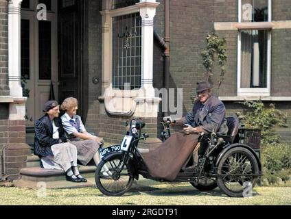 A man in a motorised bath chair in front of a house, with two women sitting on the doorstep clearly admiring his means of transport. Stock Photo