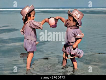 Two toddlers paddling in the sea, each one with a bucket in hand. The one on the right also carries a small watering can. Stock Photo