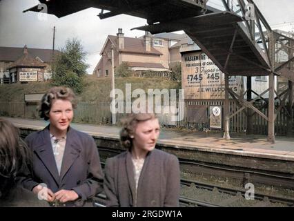 Two young women at a railway station in South London.  An advertisement on the opposite platform is for property, houses of character for ú745 freehold.  Another sign advertises self-contained flats to let. Stock Photo