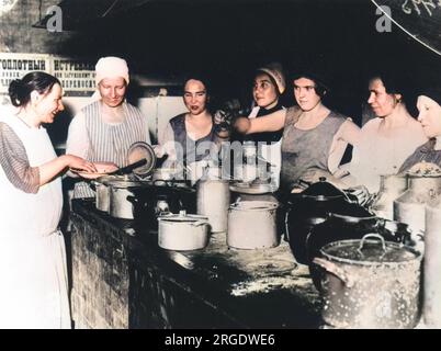 Seven women busying themselves around a stone work top adorned with an array of large tins and pots in a 1920's Russian communal kitchen. Stock Photo