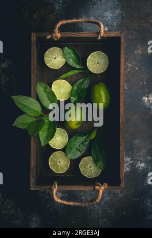 Limes, cut and whole, and Lime Leaves in wood box on dark background Stock Photo