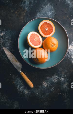 Pink grapefruit, one halved and two whole, in blue ceramic bowl with knife on dark background Stock Photo