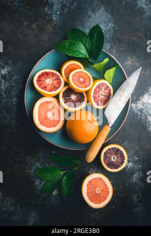 Blood Oranges and pink grapefruit, halved and whole, with citrus leaves, in blue ceramic bowl and on table, with knife on dark background Stock Photo