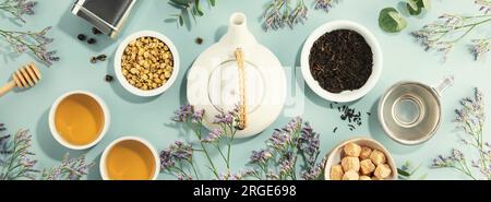 A white porcelain teapot, two cups, suga and assortment of dry tea in white ceramic bowls on blue background.  Top view. Banner Stock Photo