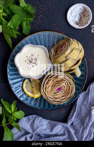 Steamed artichokes served with a bowl of dipping sauce on a dark blue plate. Stock Photo