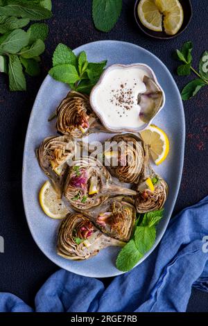 Roasted artichokes served on an oval plate with a bowl of dipping sauce on the side. Stock Photo