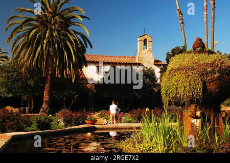 People walk within the historic flower gardens of the San Juan Capistrano Mission in California on a sunny day Stock Photo