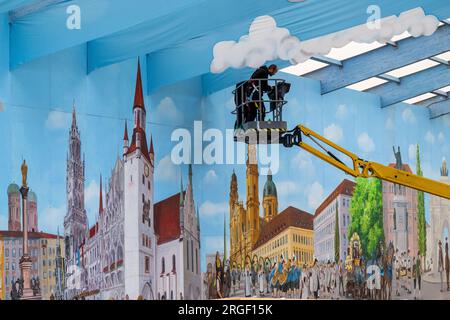 Munich, Germany. 09th Aug, 2023. Two workers on a lifting platform attach blue fabric panels with a cloud pattern under the roof structure of a beer tent on the Oktoberfest grounds. Large pictures with motifs of the Bavarian capital hang on the walls. The 188th Oktoberfest is scheduled to take place this year from Sept. 16 to Oct. 3, 2023. Credit: Peter Kneffel/dpa/Alamy Live News